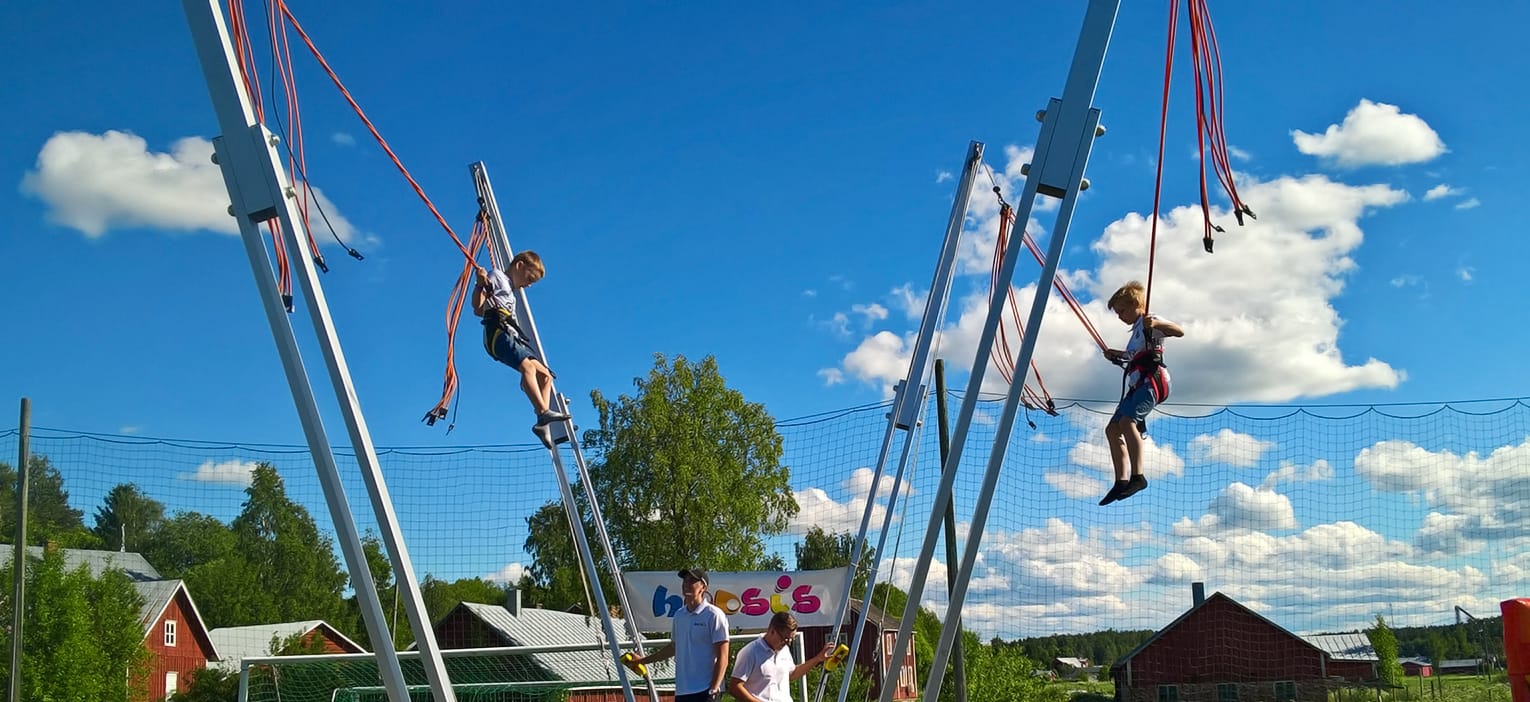 Kaksi lasta lentää korkealla bungy-trampoliinin valjaissa sinistä kesätaivasta vasten