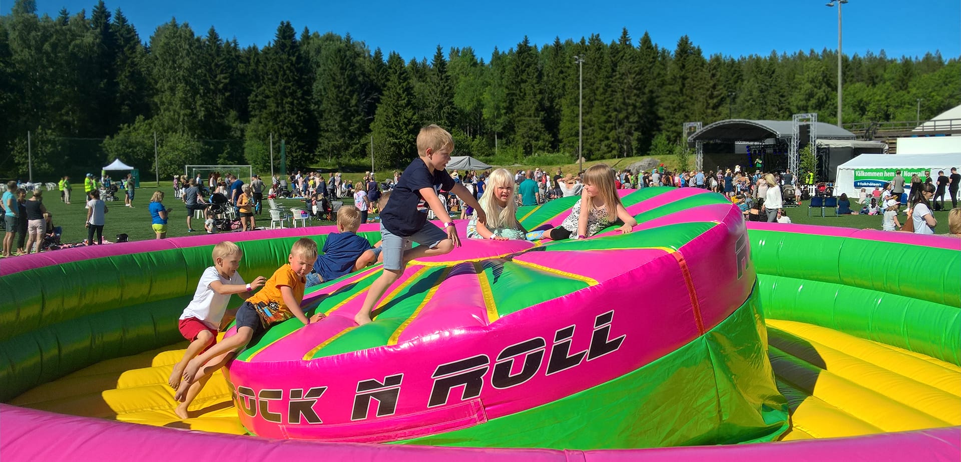 Lapsia kiipeämässä pinkille Rock n Roll -pomppulinnalle aurinkoisessa kesätapahtumassa, taustalla yleisöä ja esiintymislava